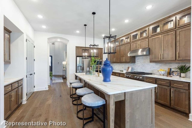 kitchen with appliances with stainless steel finishes, hardwood / wood-style flooring, decorative light fixtures, an island with sink, and light stone counters