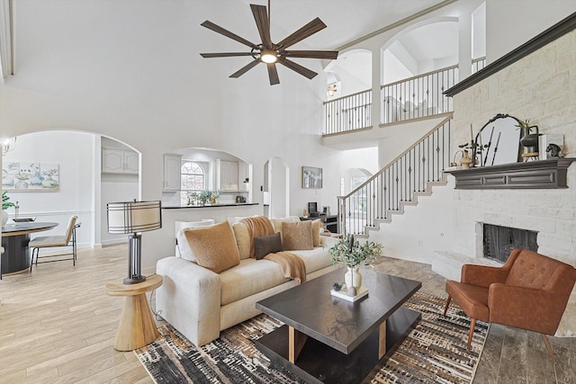 living room featuring light wood finished floors, stairs, a ceiling fan, and a stone fireplace