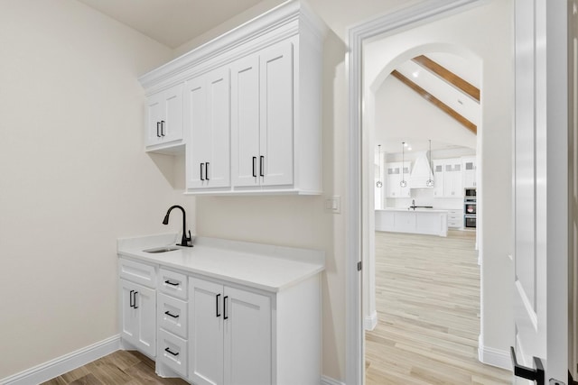 bar featuring custom range hood, white cabinets, light wood-type flooring, sink, and vaulted ceiling with beams