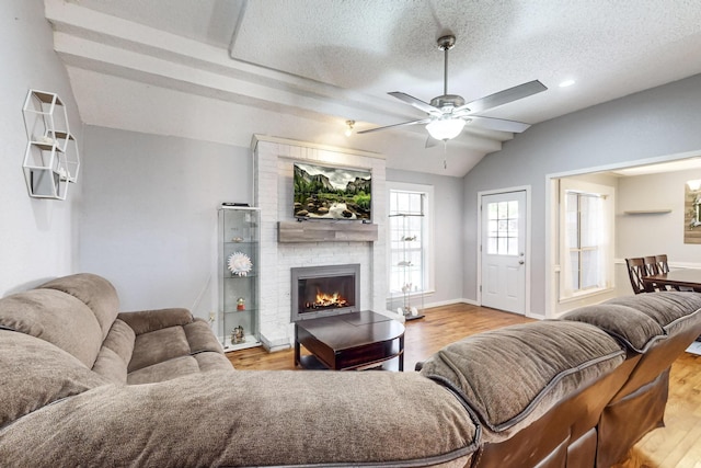 living room with a textured ceiling, a brick fireplace, light hardwood / wood-style flooring, ceiling fan, and lofted ceiling