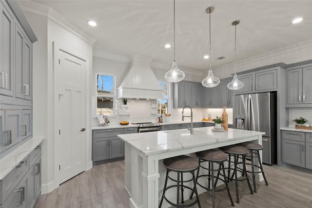 kitchen with decorative light fixtures, gray cabinets, custom range hood, stainless steel appliances, and a kitchen island with sink