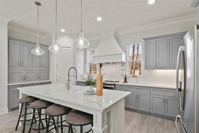 kitchen with an island with sink, hanging light fixtures, stainless steel appliances, light stone countertops, and custom range hood