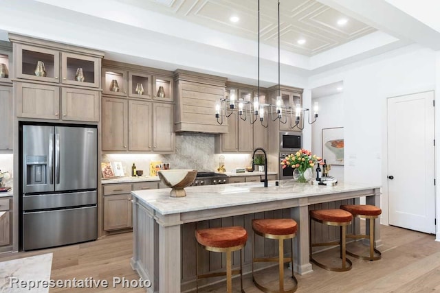 kitchen featuring appliances with stainless steel finishes, backsplash, light stone counters, a center island with sink, and coffered ceiling