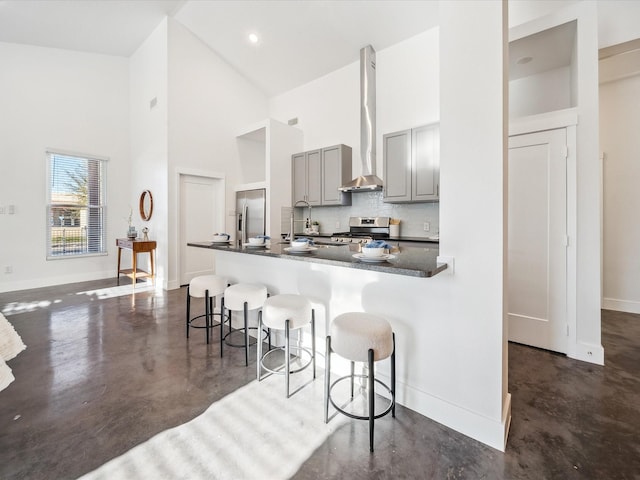 kitchen featuring a towering ceiling, a kitchen bar, gray cabinetry, stainless steel appliances, and wall chimney exhaust hood