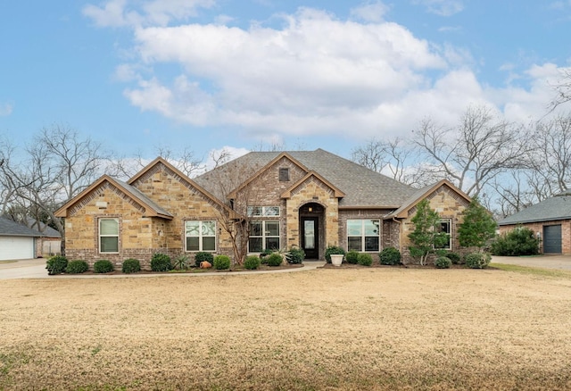 french country style house featuring stone siding, a front lawn, brick siding, and roof with shingles