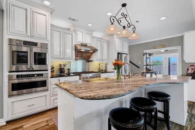 kitchen with tasteful backsplash, hanging light fixtures, white cabinets, a center island with sink, and double oven