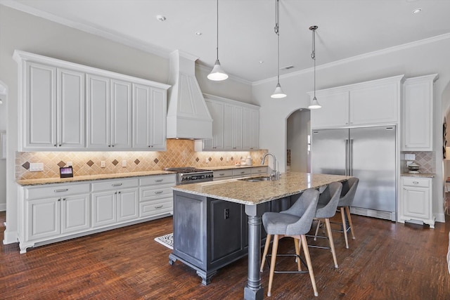 kitchen featuring appliances with stainless steel finishes, decorative light fixtures, white cabinetry, custom exhaust hood, and a kitchen island with sink