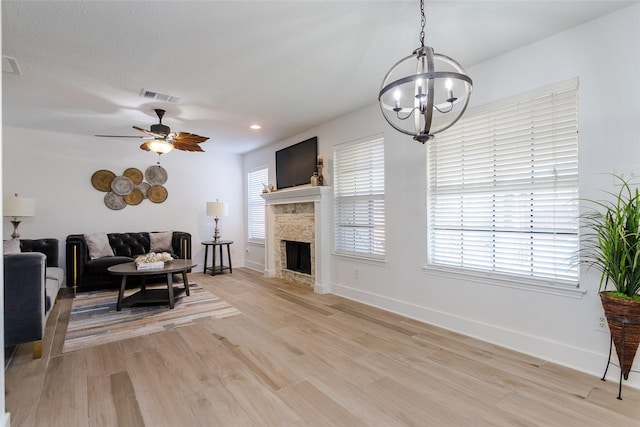 living room with ceiling fan with notable chandelier, light hardwood / wood-style flooring, and a stone fireplace