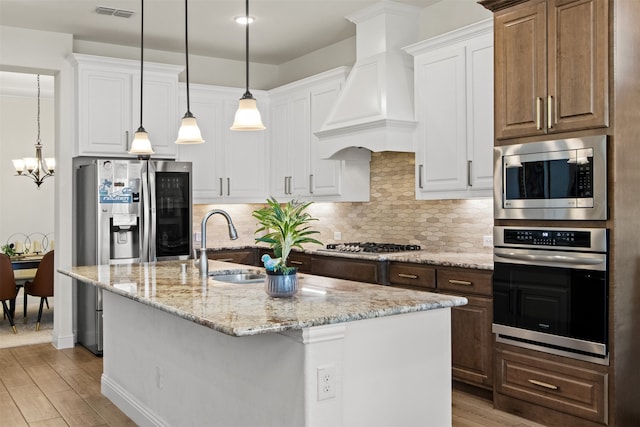 kitchen featuring custom exhaust hood, decorative light fixtures, white cabinets, an island with sink, and stainless steel appliances