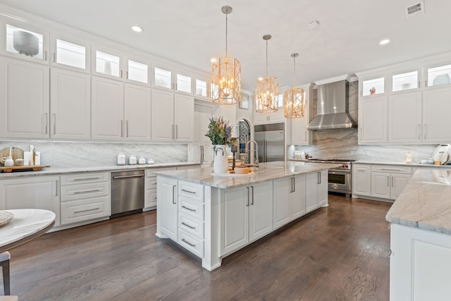 kitchen featuring hanging light fixtures, high quality appliances, white cabinets, wall chimney range hood, and an island with sink