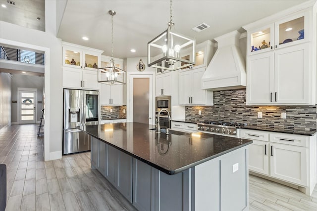 kitchen featuring an island with sink, a chandelier, custom exhaust hood, appliances with stainless steel finishes, and decorative backsplash