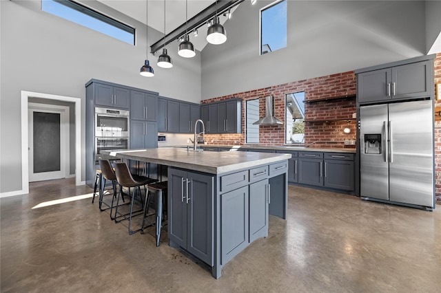 kitchen featuring appliances with stainless steel finishes, a towering ceiling, wall chimney range hood, an island with sink, and sink