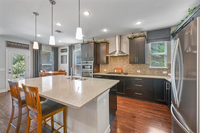 kitchen featuring appliances with stainless steel finishes, wall chimney range hood, sink, dark wood-type flooring, and an island with sink