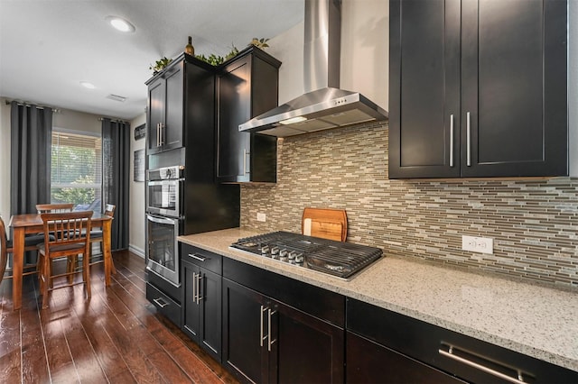 kitchen with appliances with stainless steel finishes, tasteful backsplash, wall chimney range hood, dark hardwood / wood-style flooring, and light stone counters