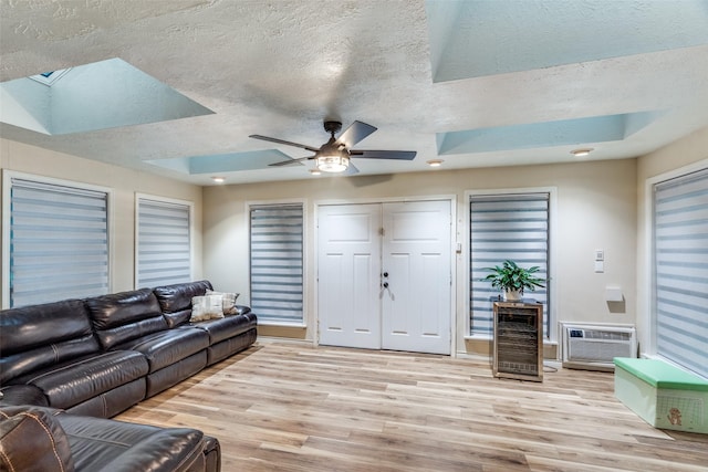 living room with a textured ceiling, an AC wall unit, light wood-type flooring, beverage cooler, and a tray ceiling