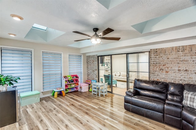 recreation room with a textured ceiling, a skylight, brick wall, light hardwood / wood-style flooring, and ceiling fan