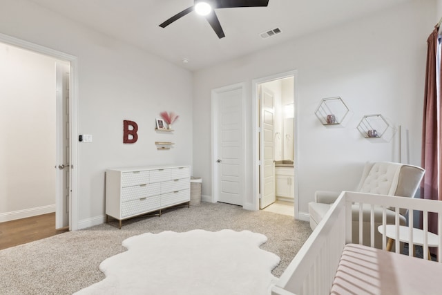 bedroom featuring light carpet, a crib, ceiling fan, and ensuite bathroom