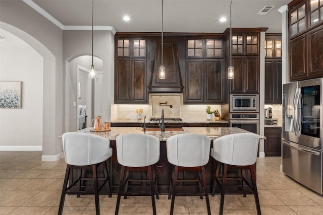 kitchen featuring dark brown cabinets, appliances with stainless steel finishes, custom exhaust hood, light tile patterned floors, and an island with sink