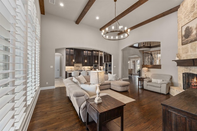 living room featuring a towering ceiling, dark wood-type flooring, a stone fireplace, beamed ceiling, and an inviting chandelier