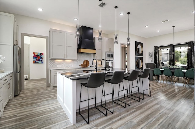 kitchen with pendant lighting, premium range hood, tasteful backsplash, a center island with sink, and stainless steel appliances