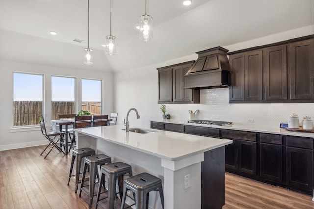 kitchen featuring custom exhaust hood, sink, hanging light fixtures, vaulted ceiling, and a center island with sink