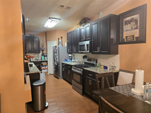 kitchen with a textured ceiling, appliances with stainless steel finishes, decorative backsplash, light stone counters, and light hardwood / wood-style flooring