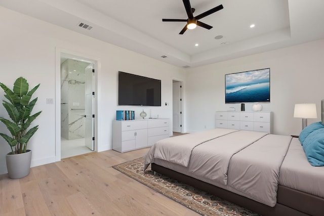 bedroom featuring a tray ceiling, ensuite bathroom, ceiling fan, and light wood-type flooring