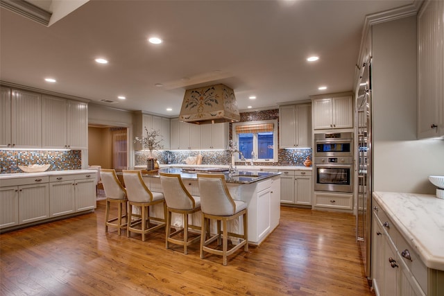 kitchen with a kitchen bar, light stone countertops, light wood-type flooring, a center island with sink, and double oven