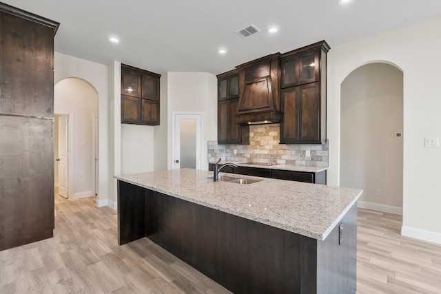 kitchen with light hardwood / wood-style floors, sink, dark brown cabinets, light stone counters, and a center island with sink