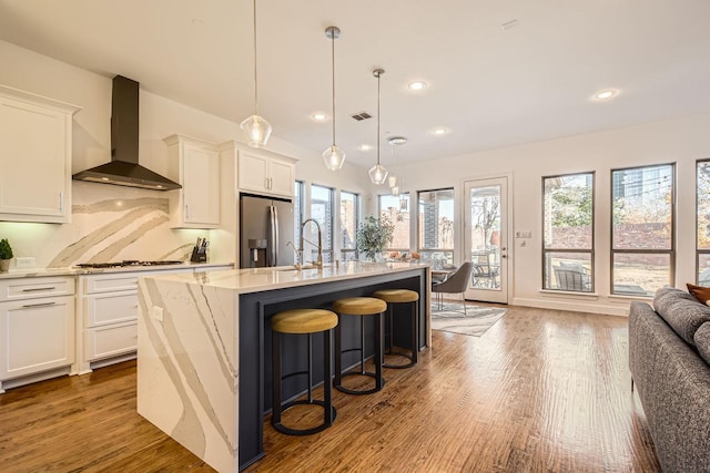 kitchen with wall chimney range hood, white cabinetry, a center island with sink, and stainless steel fridge with ice dispenser