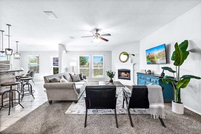 tiled living area featuring a warm lit fireplace, a textured ceiling, a ceiling fan, and baseboards