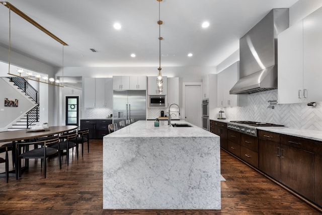 kitchen with wall chimney range hood, built in appliances, an island with sink, sink, and hanging light fixtures