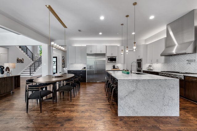 kitchen with decorative light fixtures, dark wood-type flooring, wall chimney range hood, a center island with sink, and built in appliances