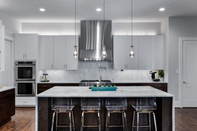 kitchen featuring wall chimney range hood, a breakfast bar, light stone counters, a center island with sink, and double oven