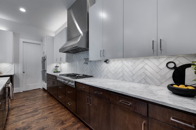 kitchen featuring wall chimney exhaust hood, white cabinetry, stainless steel appliances, decorative backsplash, and light stone counters