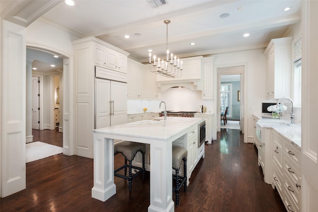 kitchen with white cabinetry, beam ceiling, pendant lighting, light stone counters, and a center island with sink