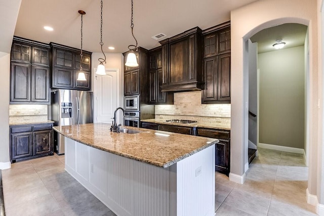 kitchen featuring appliances with stainless steel finishes, wall chimney range hood, dark brown cabinets, an island with sink, and light stone counters