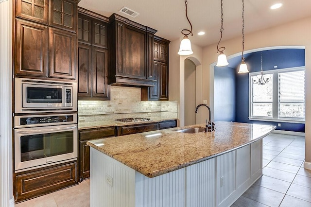 kitchen featuring dark brown cabinets, appliances with stainless steel finishes, sink, light stone counters, and a center island with sink