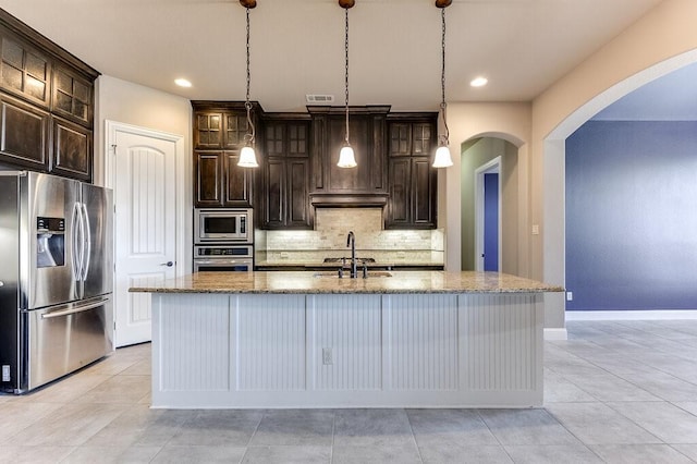 kitchen with dark brown cabinets, stainless steel appliances, a center island with sink, and decorative light fixtures