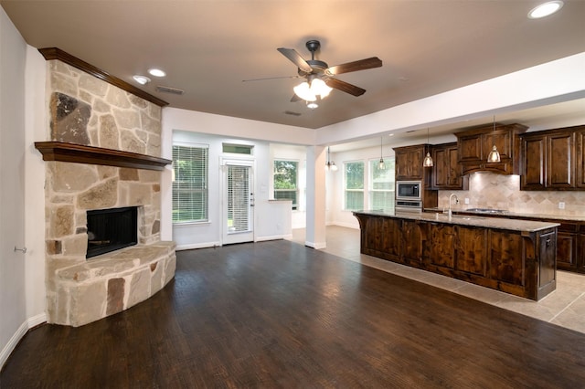 kitchen with a stone fireplace, appliances with stainless steel finishes, pendant lighting, backsplash, and an island with sink