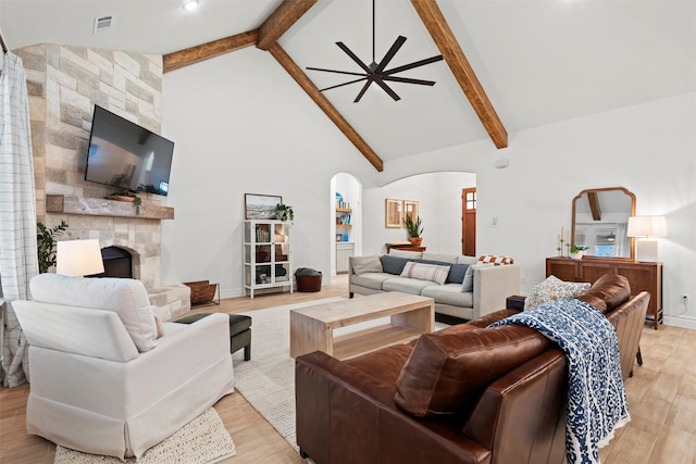 living room with a stone fireplace, light wood-type flooring, high vaulted ceiling, beam ceiling, and ceiling fan