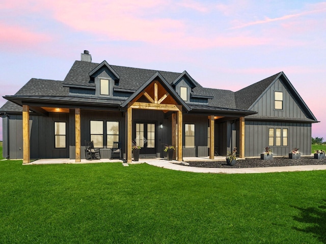 back of house at dusk with french doors, roof with shingles, a lawn, board and batten siding, and a chimney