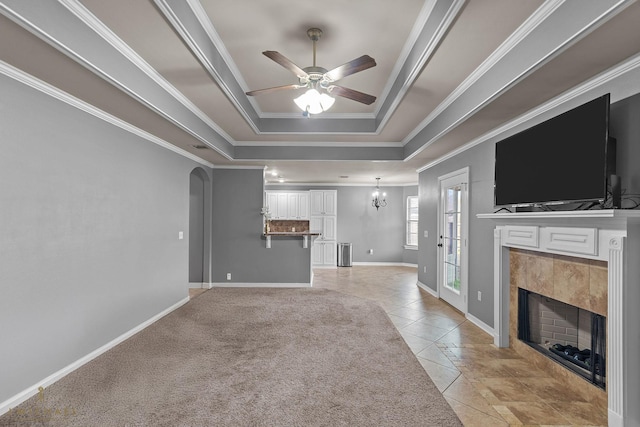 unfurnished living room featuring a tile fireplace, ceiling fan with notable chandelier, light tile patterned floors, a tray ceiling, and crown molding