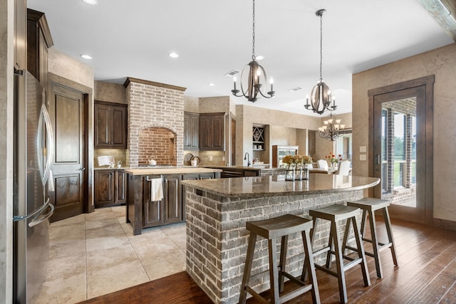 kitchen with freestanding refrigerator, a breakfast bar, dark brown cabinets, a center island with sink, and a notable chandelier