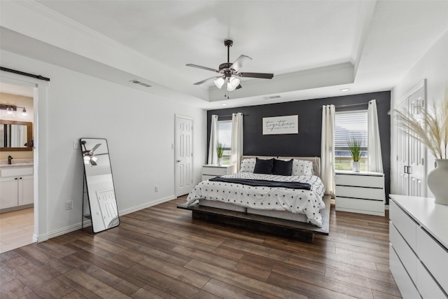 bedroom with dark hardwood / wood-style floors, connected bathroom, ceiling fan, a tray ceiling, and a barn door