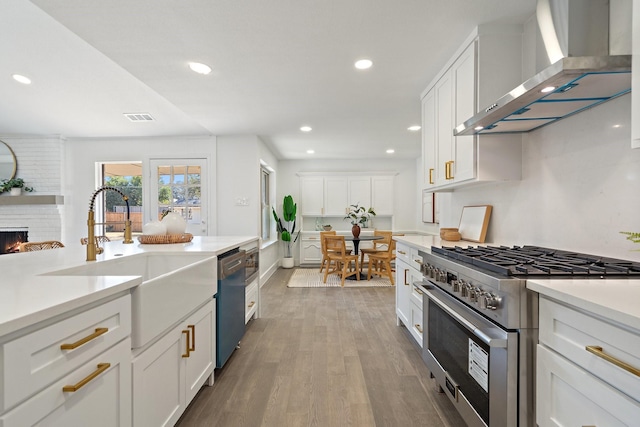 kitchen with appliances with stainless steel finishes, sink, white cabinetry, a brick fireplace, and wall chimney range hood