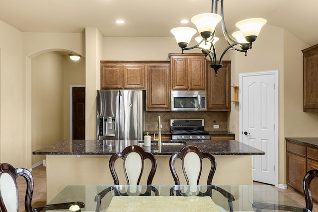 kitchen with dark stone countertops, appliances with stainless steel finishes, decorative light fixtures, backsplash, and an island with sink