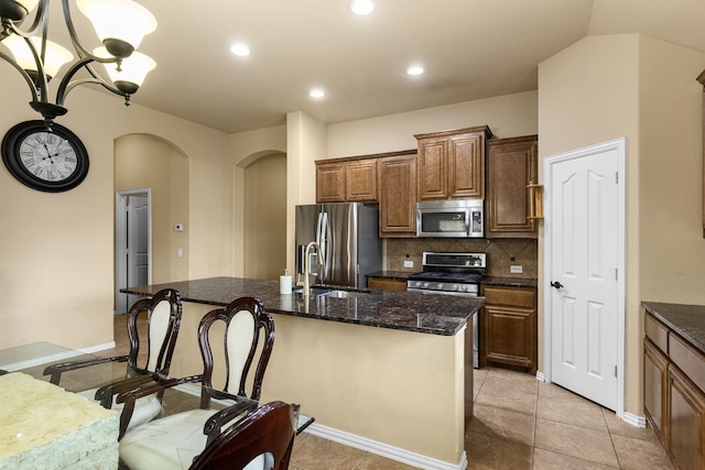 kitchen with appliances with stainless steel finishes, dark stone countertops, decorative light fixtures, backsplash, and an island with sink