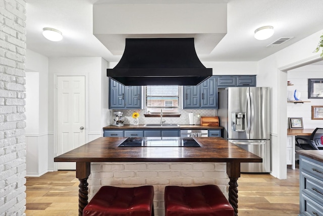 kitchen featuring butcher block counters, range hood, stainless steel refrigerator with ice dispenser, black electric cooktop, and sink