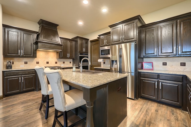 kitchen featuring appliances with stainless steel finishes, wood-type flooring, an island with sink, premium range hood, and sink